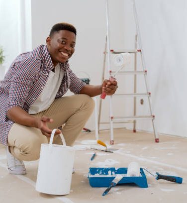 A smiling young boy in brown jeans and a red plaid shirt holds a roller and picks up a can of white paint. He is getting ready to painting the walls of an apartment he is renovating